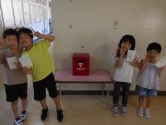 赤い小さな手作りのポストの前ではがきを持ちピースサインの子ども4人