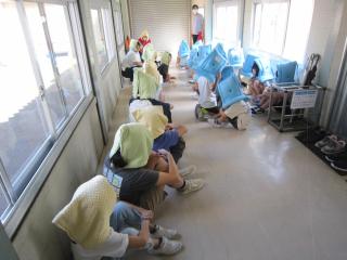 防災頭巾をかぶり廊下に並んで座っている子供たち