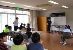 ホールのホワイトボード前で話す講師と熱心に聞く子どもたち