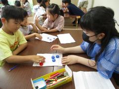 白い紙の上に青と赤のペットボトルキャップを並べて対戦する子ども達